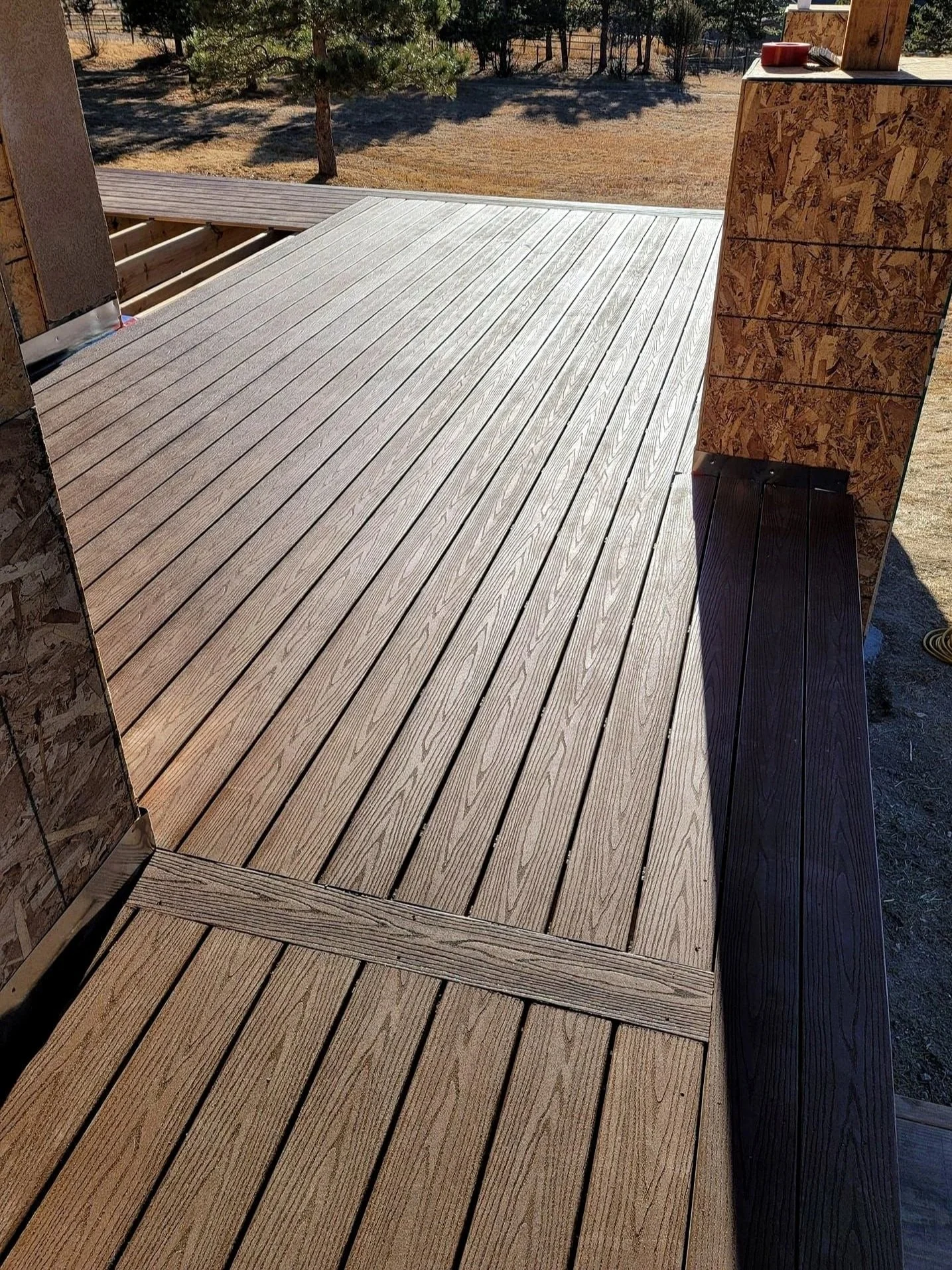Wooden deck with newly installed grayish wood planks, part of a porch or balcony, with a background of a yard with trees and dry grass.