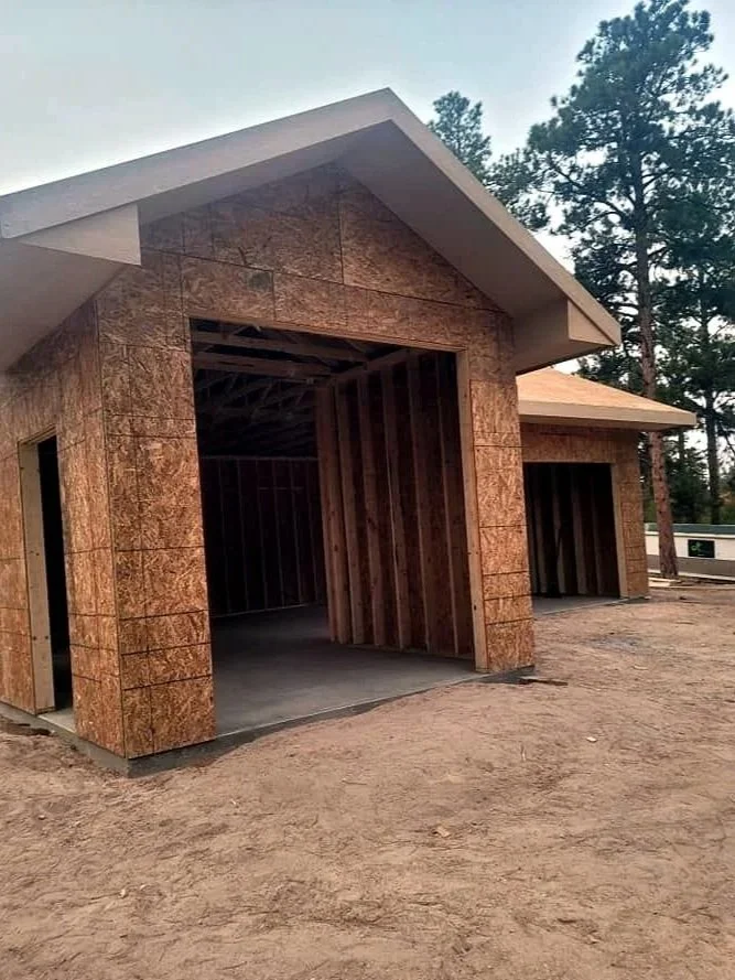 A house under construction with exposed wood framing and plywood sheathing on the exterior, and a sloped roof.