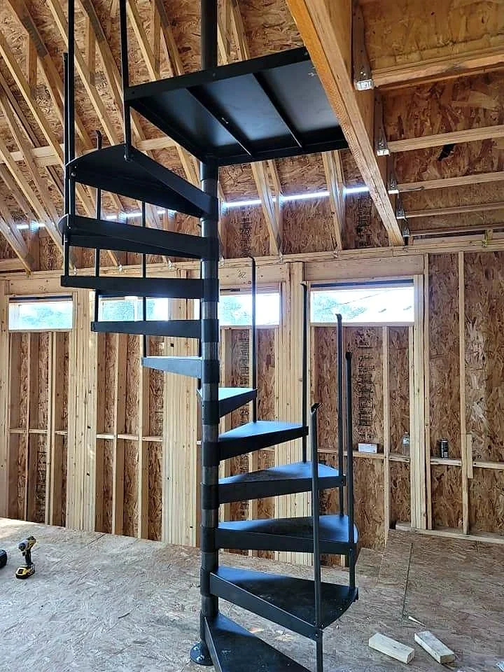 Interior of a building under construction featuring a black spiral staircase with open risers and a black landing at the top, surrounded by unfinished wooden framing and OSB wall sheathing.