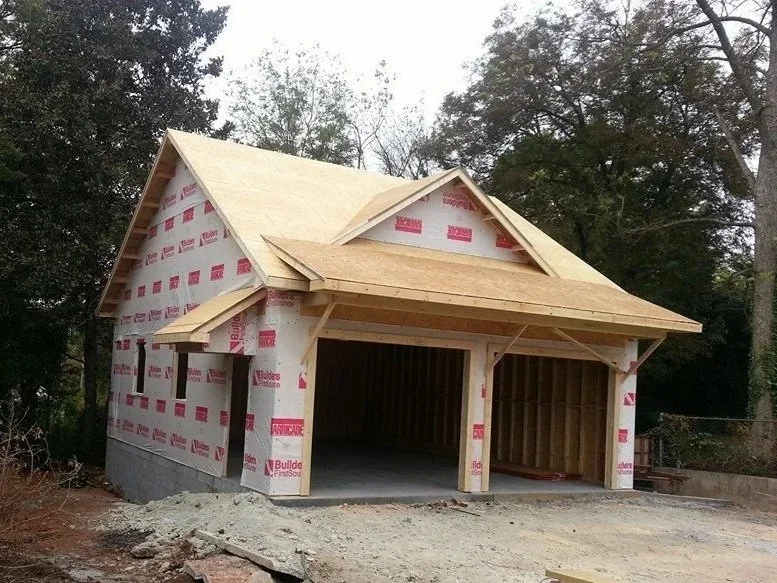 A house under construction with wooden framing and wrapped in building wrap, situated outdoors among trees.