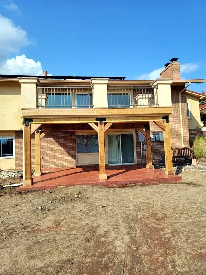 Backyard of a house featuring a newly built wooden deck with support beams, plywood flooring, and a railing on the upper level, with sliding glass doors and windows beneath.