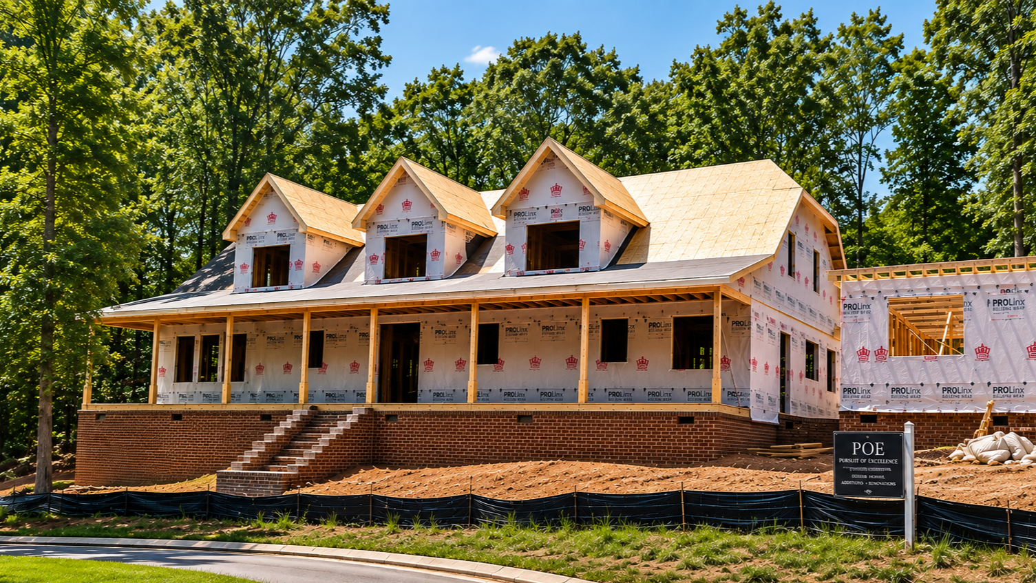 A house under construction with a brick foundation, wooden framing, and a partially completed roof, surrounded by trees and a clear blue sky.