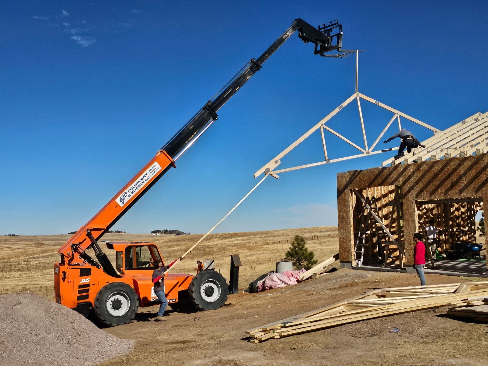 Construction site with a crane lifting a roof truss onto a building under construction. Workers are present, some on the ground and one on the roof.