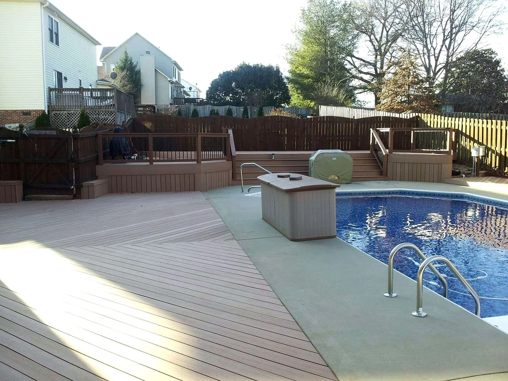 Backyard with outdoor pool, wooden deck, stairs, fence, and trees in the background.