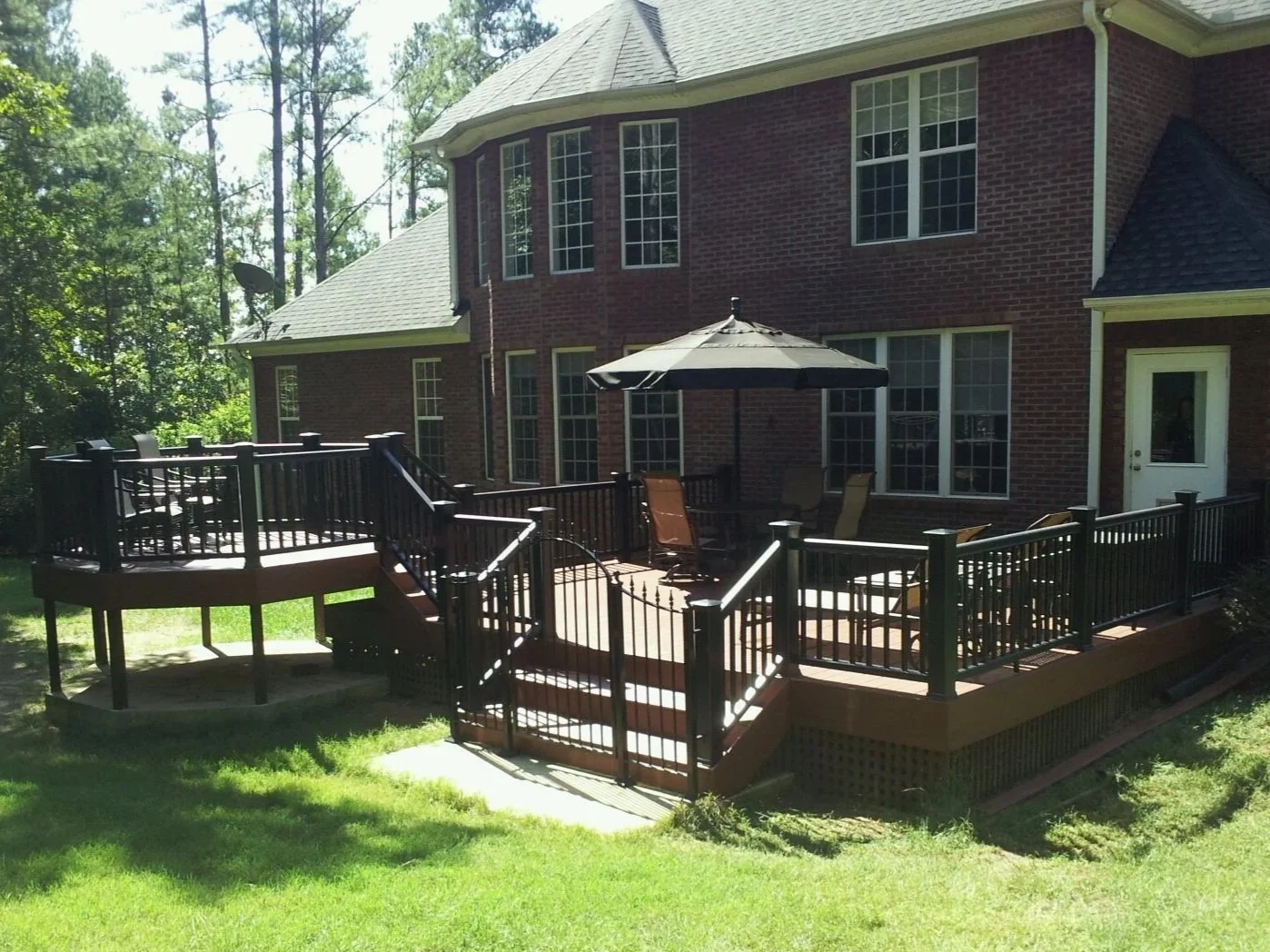 Backyard deck with patio furniture, umbrella, and railing attached to a brick house with multiple windows surrounded by trees.