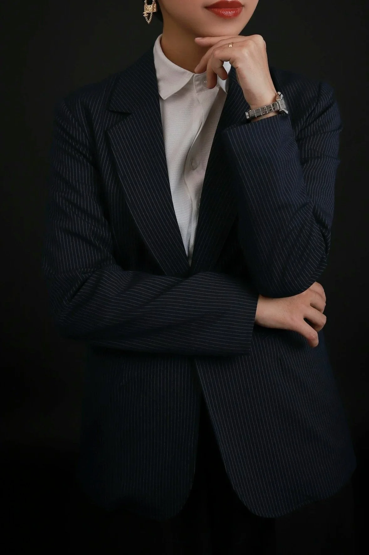 A woman in formal business attire, wearing a pinstripe suit, white shirt, and accessories, with her hand resting on her chin, against a black background.