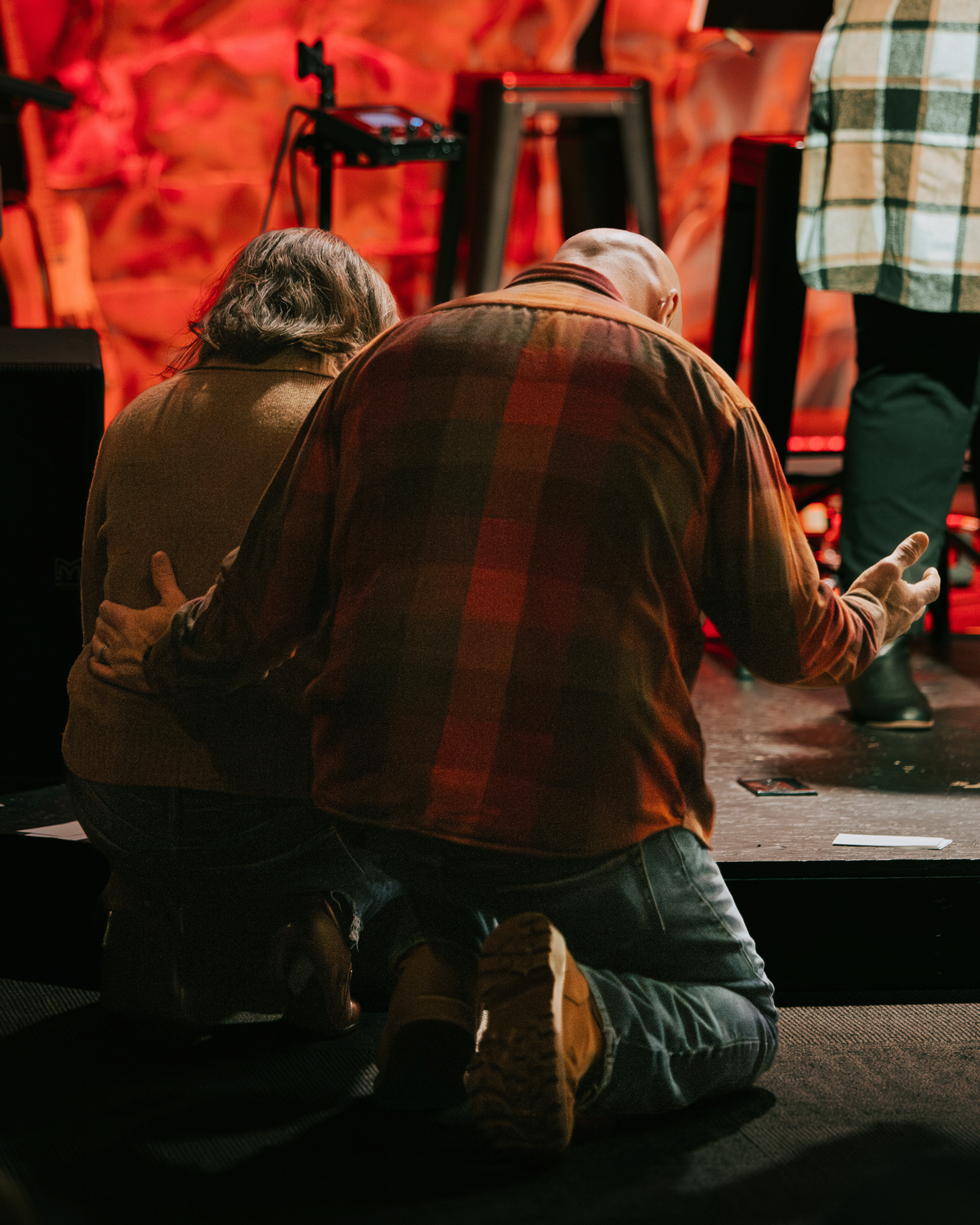 Two people kneeling on the floor and praying backstage at a performance, with stage equipment and a red background.