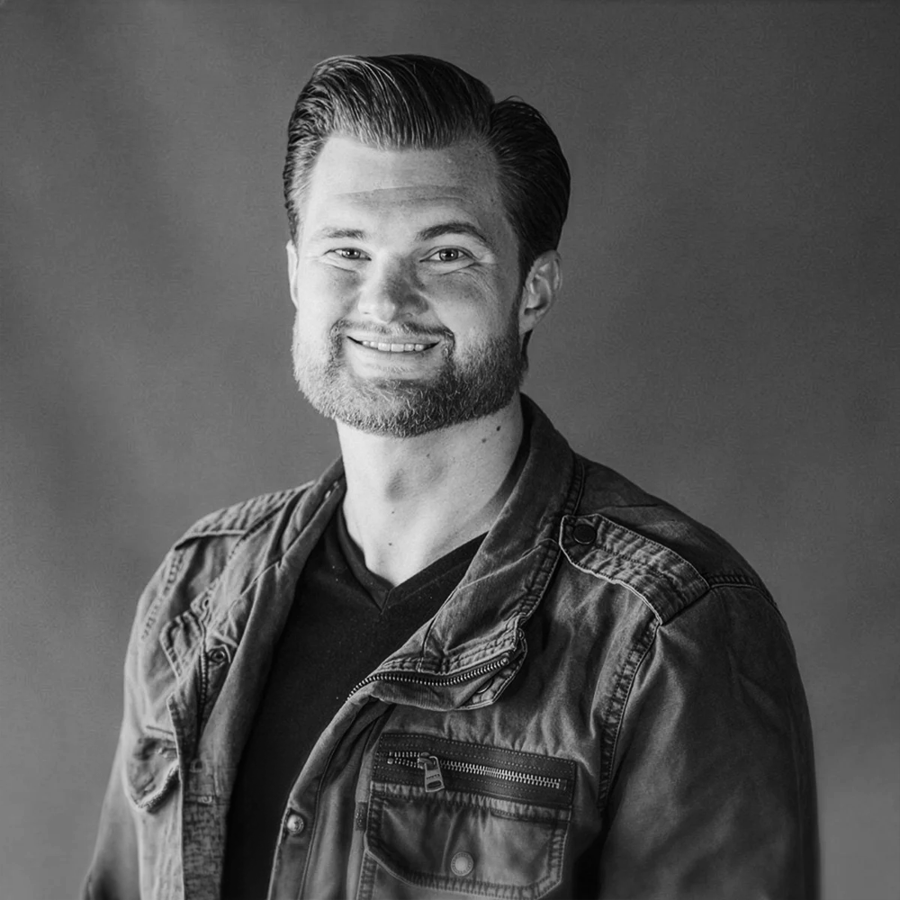 A black and white portrait of a smiling man with a beard and slicked-back hair, wearing a denim jacket over a dark shirt, against a plain background.
