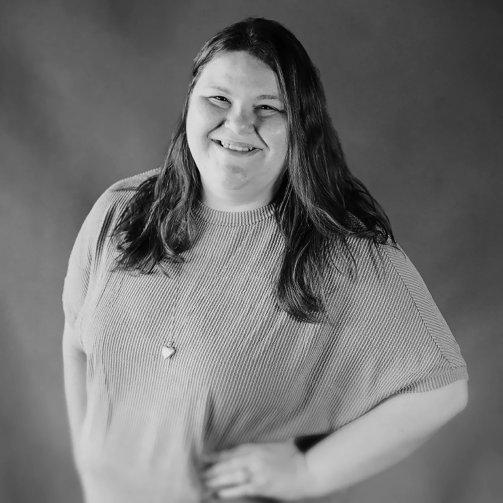 Woman with long dark hair smiling at camera, wearing a striped top and necklace, in black and white studio portrait.