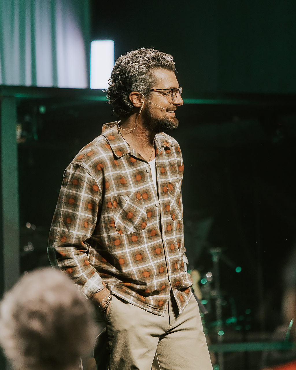 A man with curly hair and glasses, wearing a patterned shirt and beige pants, smiling while speaking on stage with dark background.