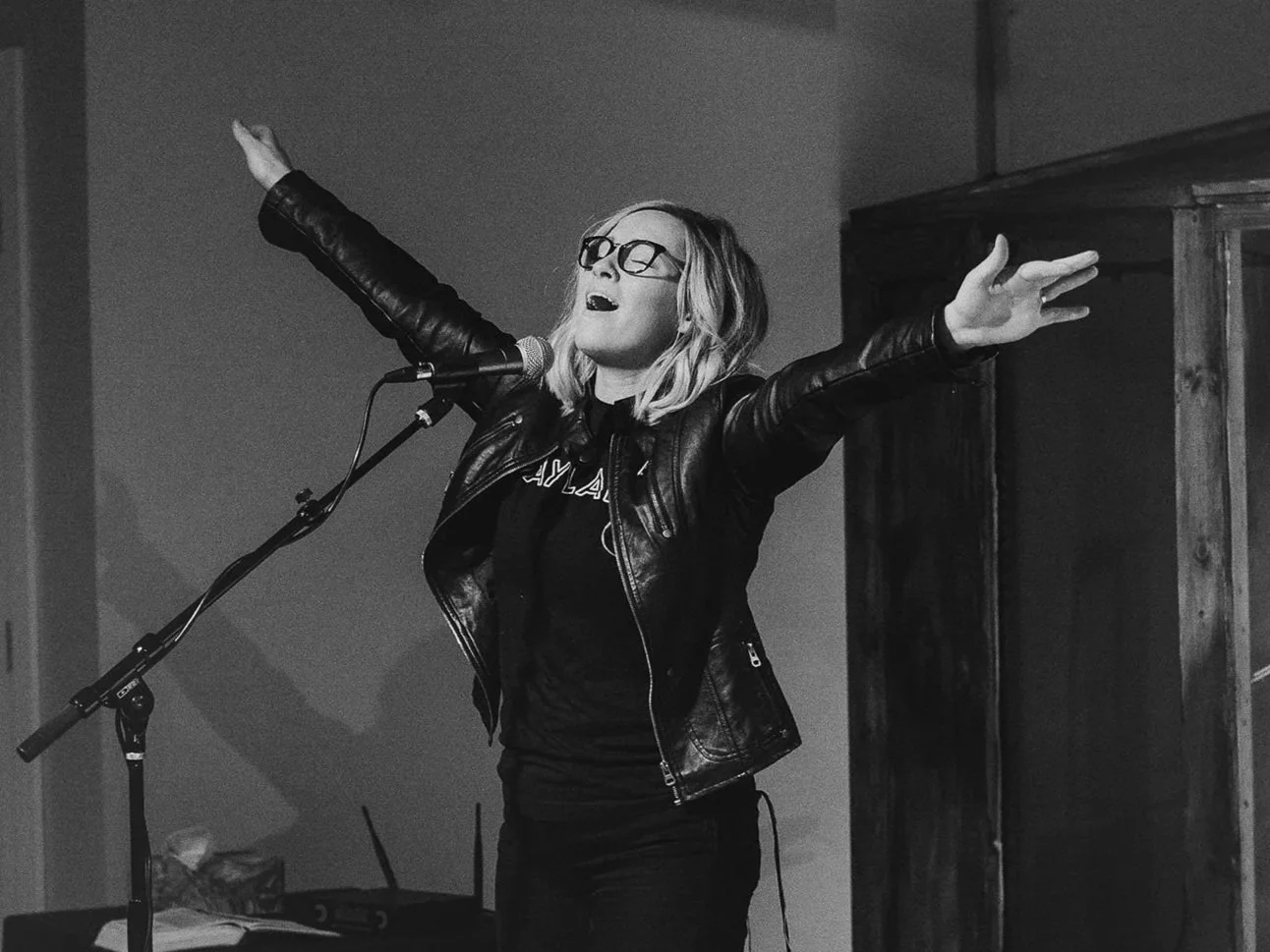 A woman wearing glasses and a leather jacket singing into a microphone with her arms raised in a performance setting.