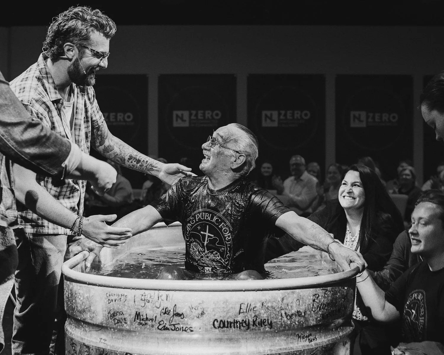 A man with glasses and tattoos is smiling and being baptized in a large water basin, with others smiling and reaching out to him. The background shows an audience and signs that say "NL Zero."