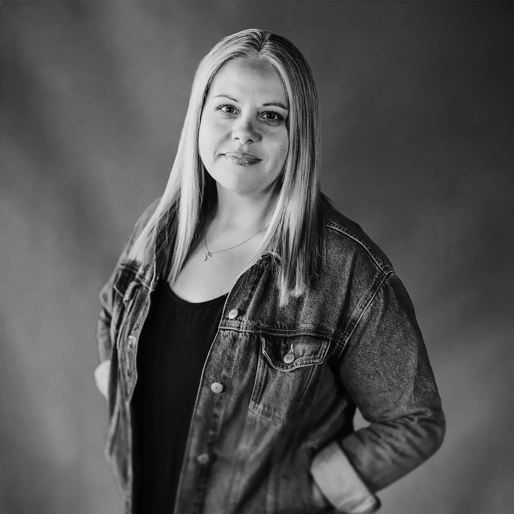 Black and white portrait of a woman with shoulder-length straight hair, wearing a denim jacket over a black top, smiling slightly, standing with one hand in her pocket against a plain background.