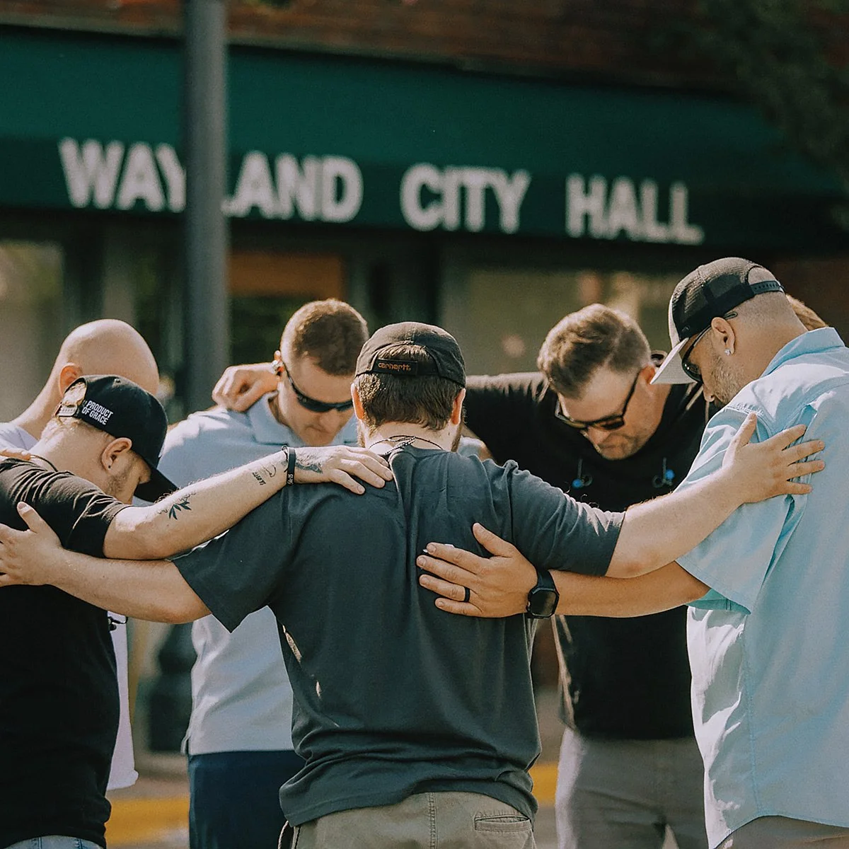 A group of people standing in a circle with their heads bowed and arms around each other, praying or showing solidarity outdoors in front of a green awning with the words 'WAY AND CITY HALL'.