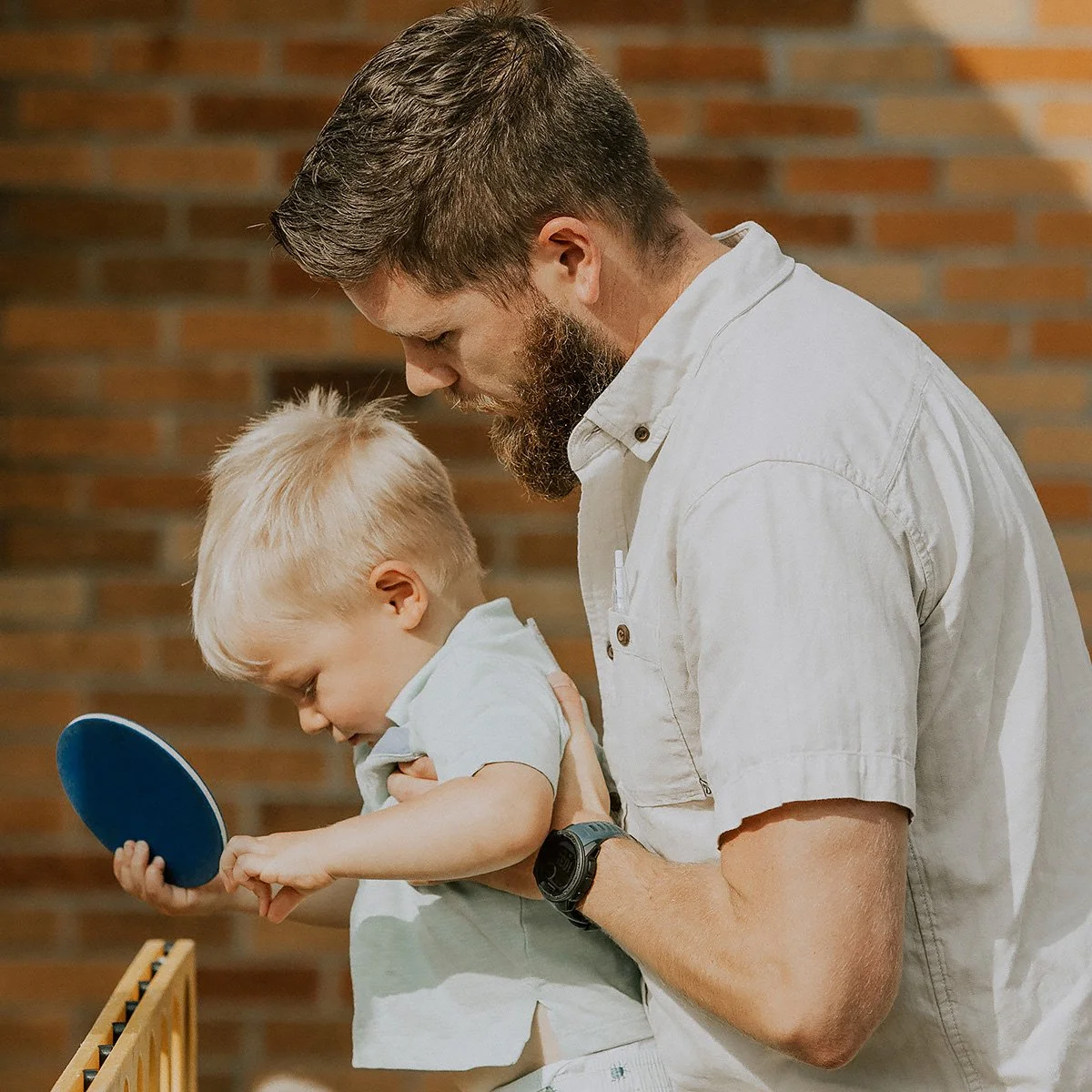 A bearded man helping a young blonde boy hold a ping pong paddle in a room with a brick wall.