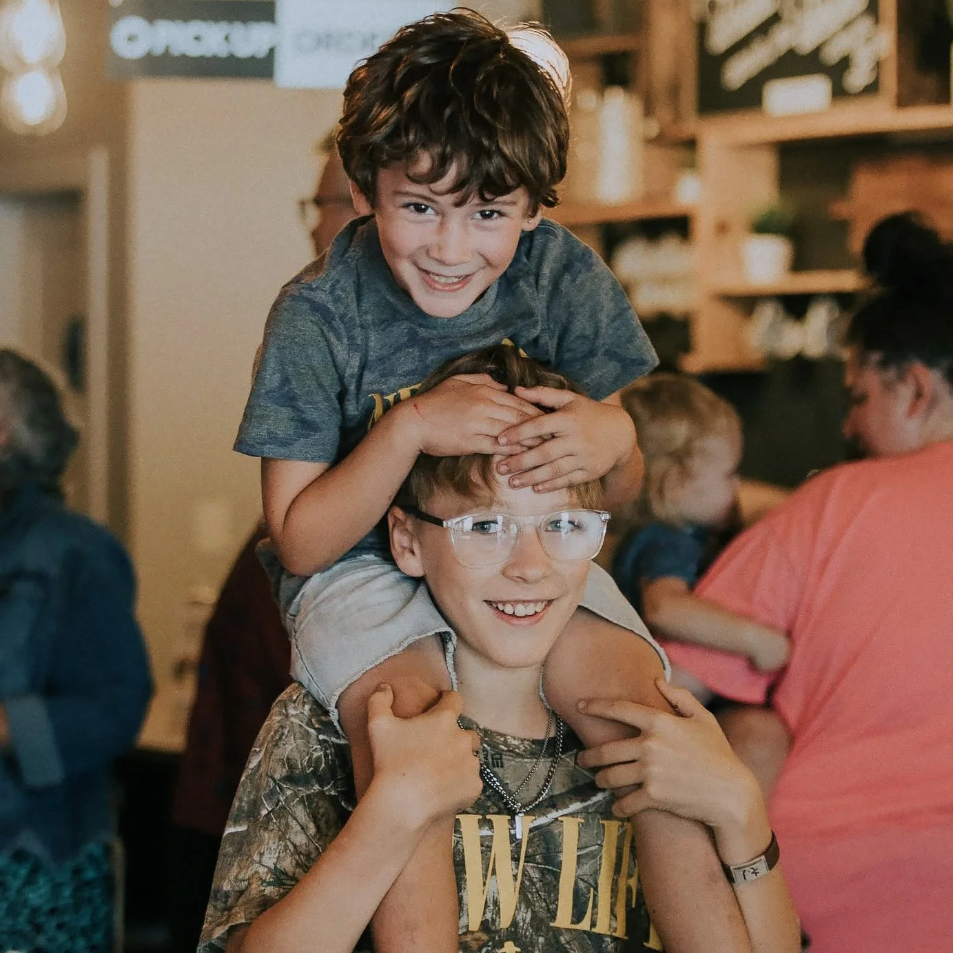 A boy with curly brown hair smiles while sitting on an adult's shoulders, with another child with glasses smiling underneath. People are in the background in an indoor setting.