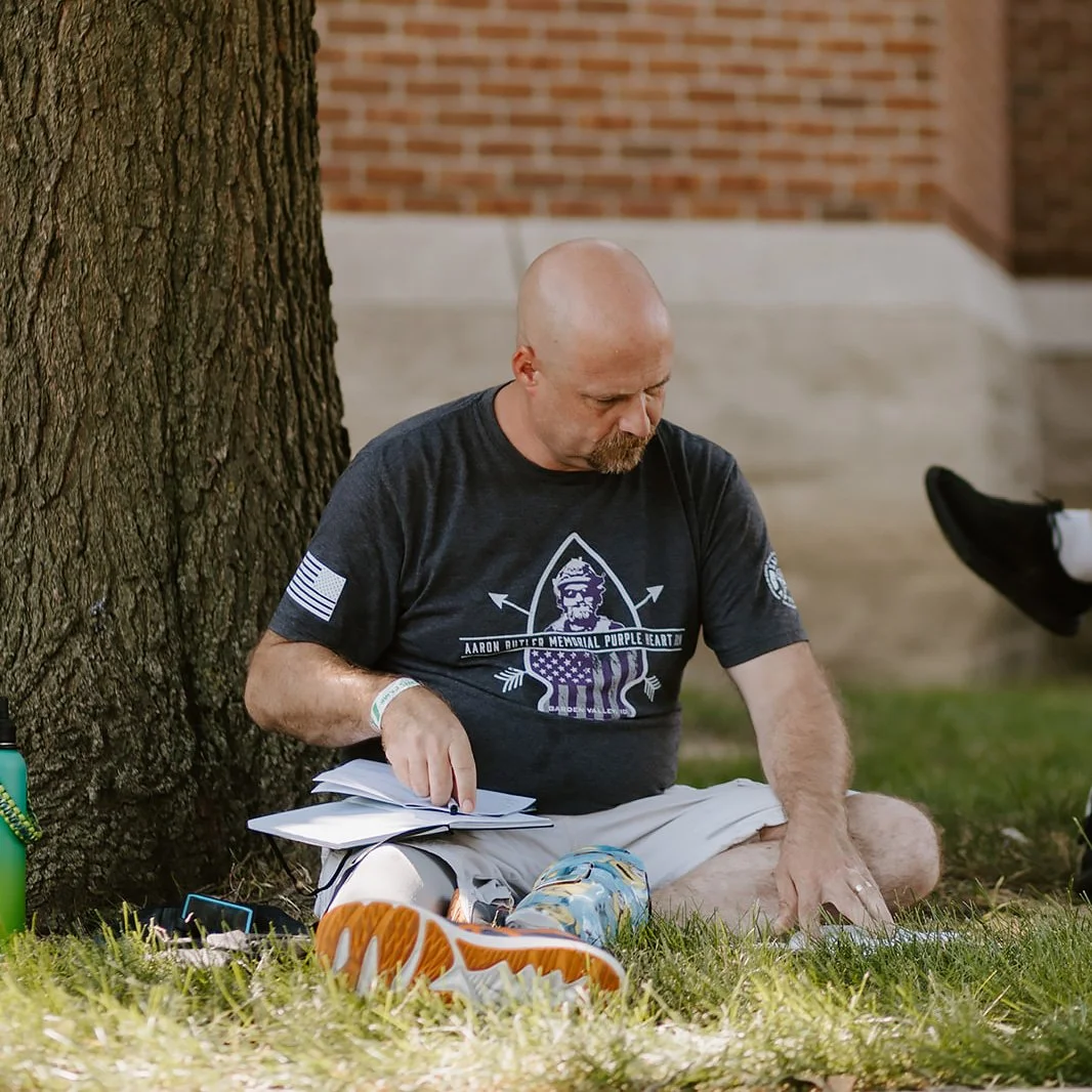 A bald man with a beard sitting on the grass next to a tree, reading a notebook. He wears a black T-shirt with a graphic design, white shorts, and orange sneakers.