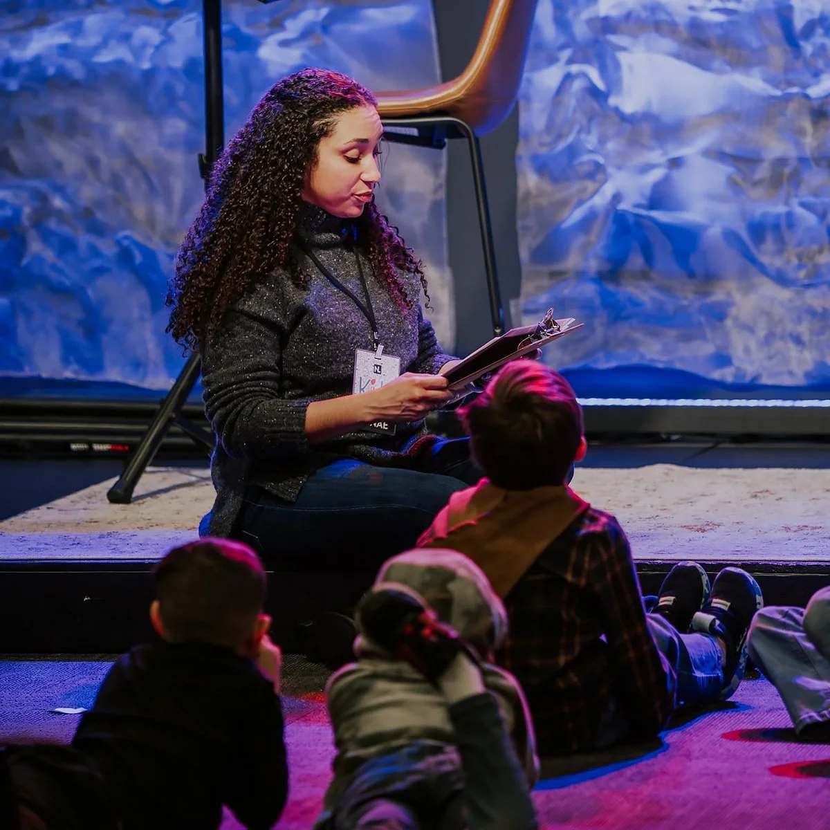 Woman with curly hair and a lanyard reading to children sitting on the floor, in front of a backdrop resembling a mountain scene.