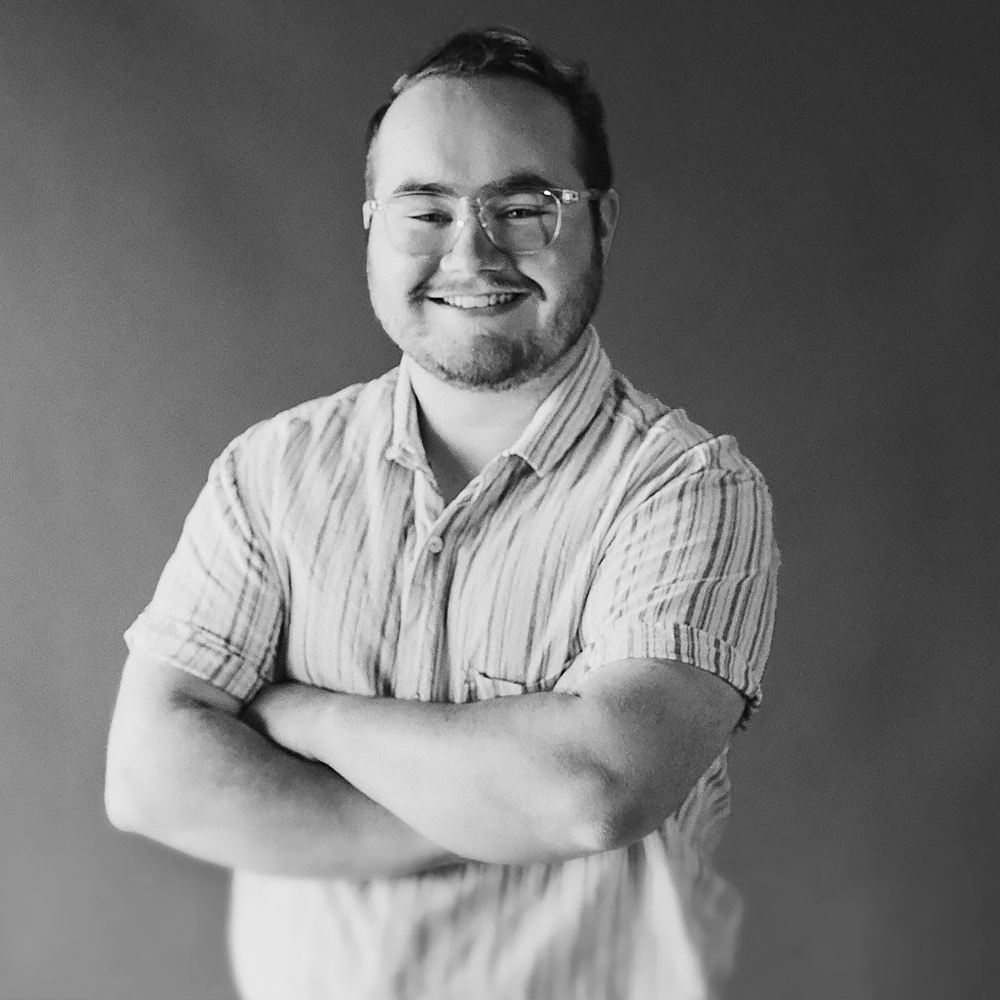 A smiling man with glasses, a beard, and short hair, wearing a striped short-sleeve shirt and standing with arms crossed against a plain background.