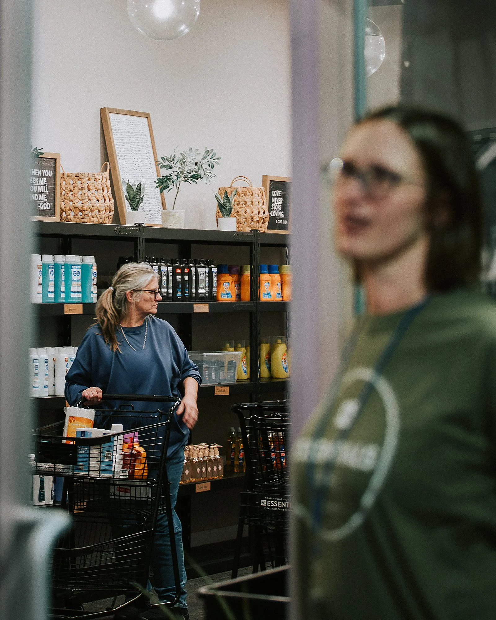 A woman with glasses and a ponytail shopping with a cart in a grocery store, seen through a gap in a shopping cart or a similar structure. Shelves behind her display household or personal care products, with some decorative items and framed signs on top.