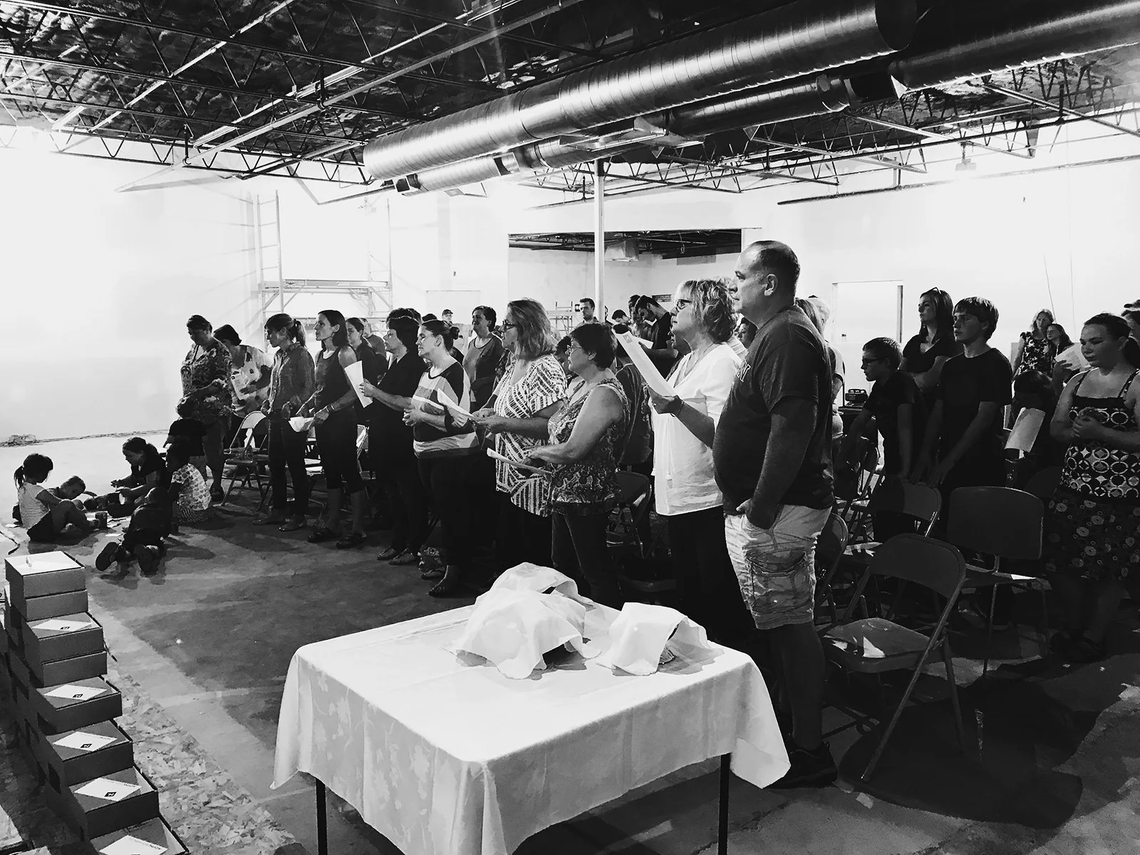 Group of people standing in line holding sheets of paper inside a large industrial space, with children sitting on the floor and boxes on a table in the foreground.