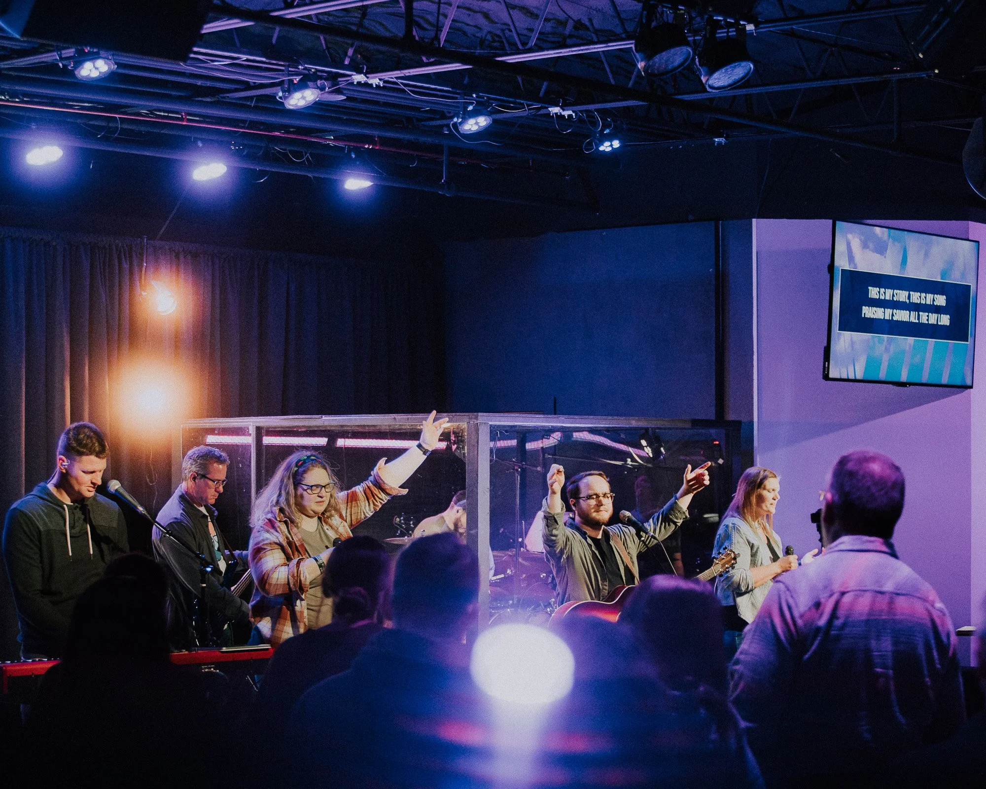 A group of people singing and playing guitars during a religious event or worship service in a darkened room with purple and blue lighting, with a screen displaying lyrics in the background.