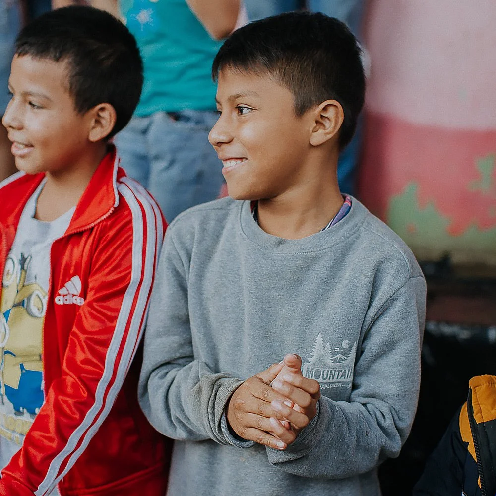 Two smiling boys standing together, one in a red Adidas jacket, the other in a gray sweatshirt with mountains illustration, in a colorful indoor setting.