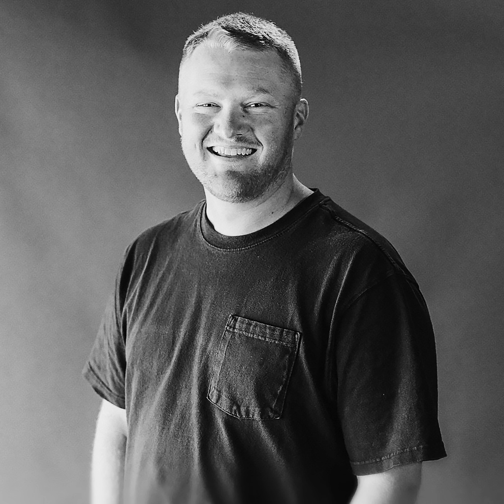 Black and white portrait of a smiling man with short hair and a beard, wearing a casual black T-shirt with a pocket, against a plain background.