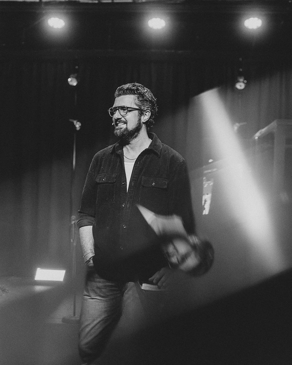 Black and white photo of a smiling man with glasses, a beard, and wavy hair, standing on stage with stage lights overhead, wearing a dark button-up shirt and jeans.