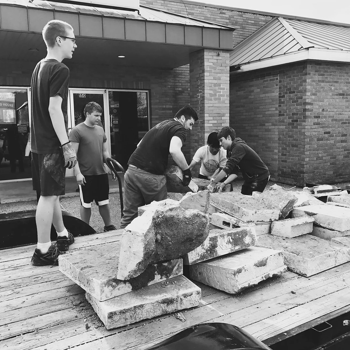 Group of five young men working together to move and arrange large stones and bricks outside a brick building, possibly during a construction or community project.
