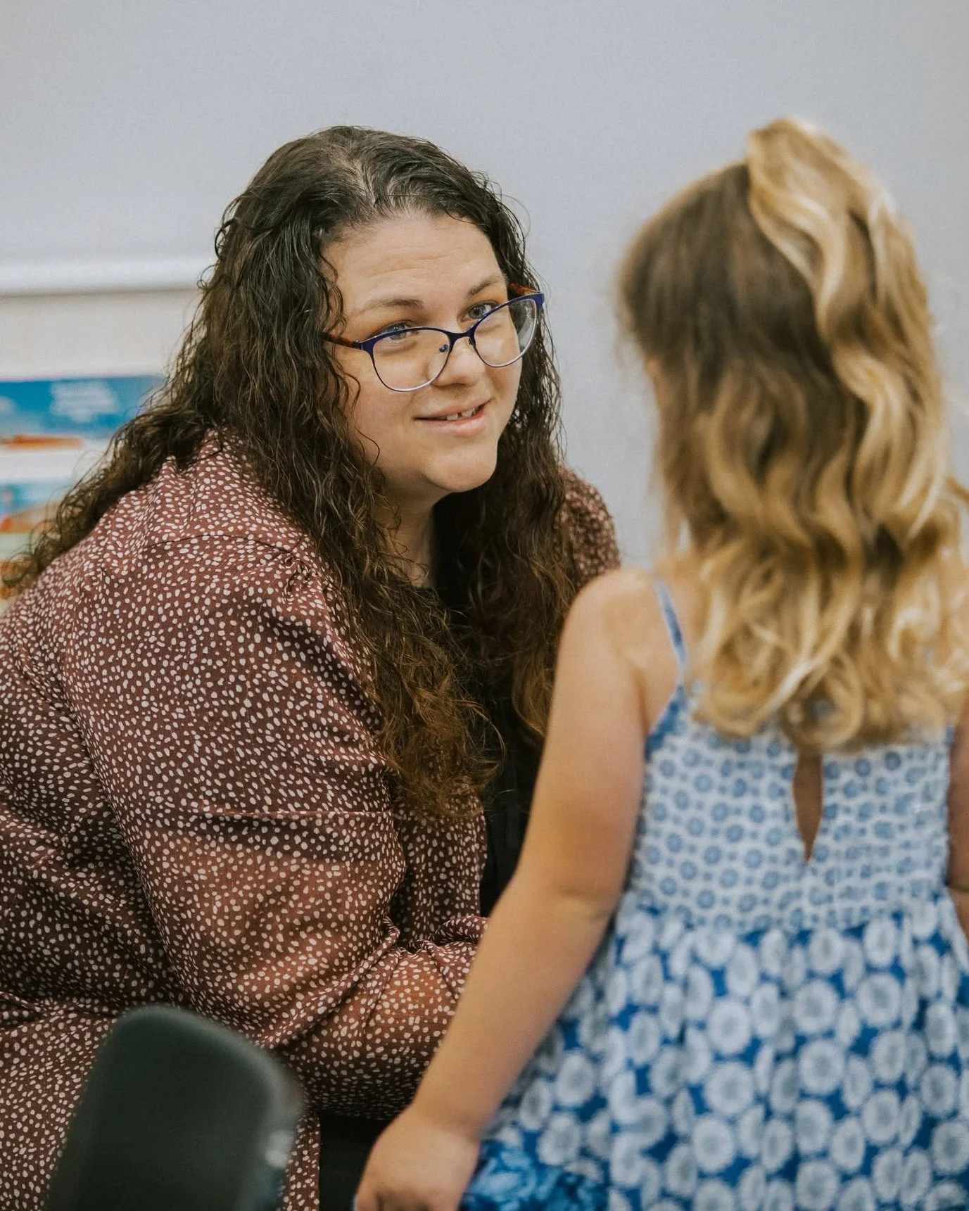 A woman with long curly brown hair wearing glasses and a brown patterned jacket smiling while talking to a young girl with long curly blonde hair in a blue floral dress.