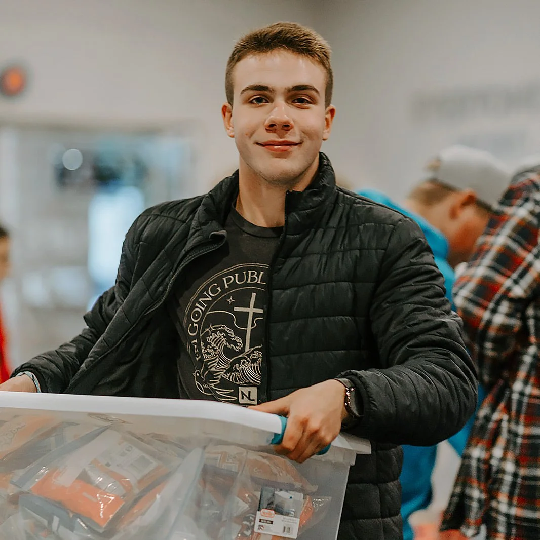 A young man with short brown hair, wearing a black quilted jacket and a black T-shirt, is smiling and holding a clear plastic container filled with food. He is in an indoor setting with blurred people in the background.
