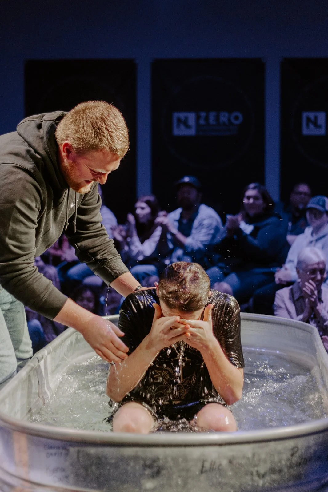 A person being baptized in a water baptismal pool with another person assisting, surrounded by an attentive audience.