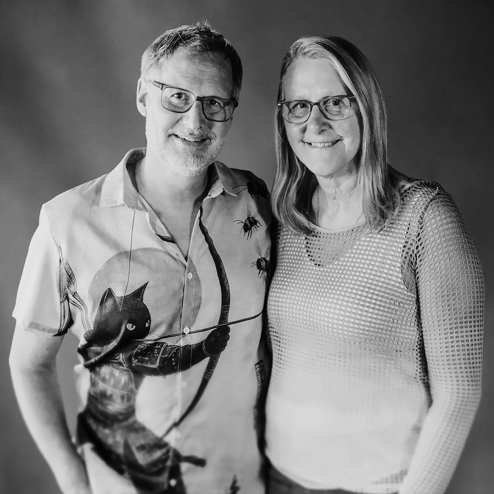 A black and white photo of a middle-aged man and woman, both wearing glasses and smiling. The man has short hair, a beard, and is wearing a short-sleeved shirt with a graphic of a black cat and a bow and arrow. The woman has shoulder-length hair and is wearing a sheer, patterned top.