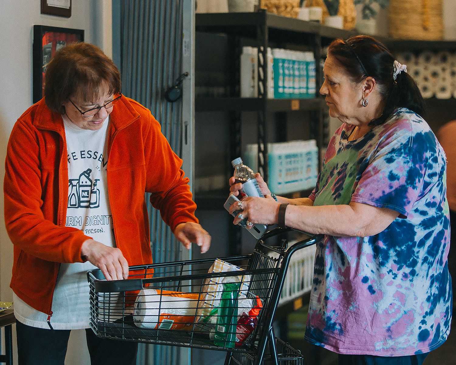 Two women are grocery shopping, one is placing items into a shopping cart while the other is holding a water bottle and a credit card. They are inside a store with shelves in the background.