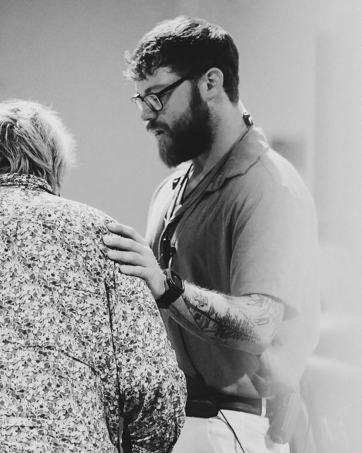A man with a beard and glasses wearing a watch and a striped shirt, placing his hand on the shoulder of an older woman with curly hair wearing a patterned shirt, in a grayscale photograph.