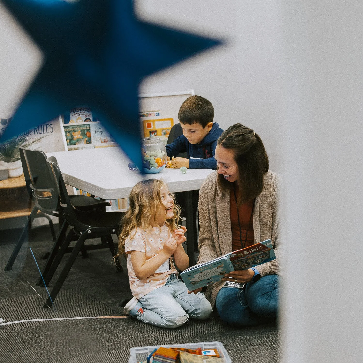 A woman and a young girl are sitting on the floor, engaged in reading a book together. A boy in the background is sitting at a table, looking down at objects on the table.