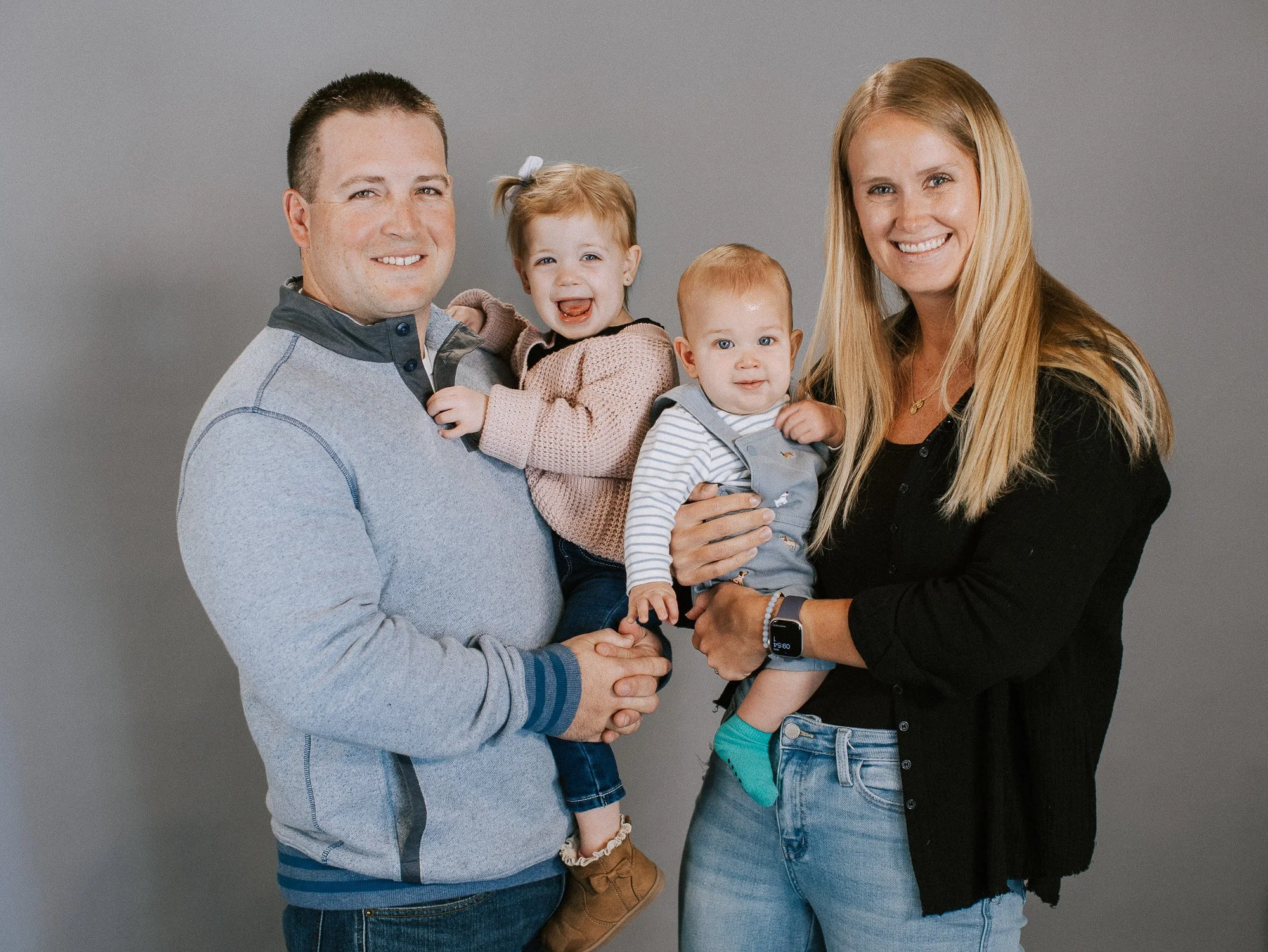 A happy family portrait with two parents and their two young children against a neutral gray background. The father is holding a young girl, and the mother is holding a baby boy. All family members are smiling and looking at the camera.