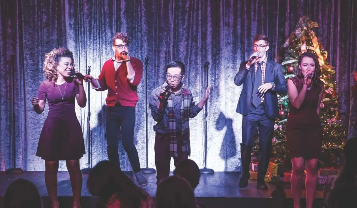 Five people performing on stage with microphones, a Christmas tree with ornaments and ribbon, and purple curtains in the background.