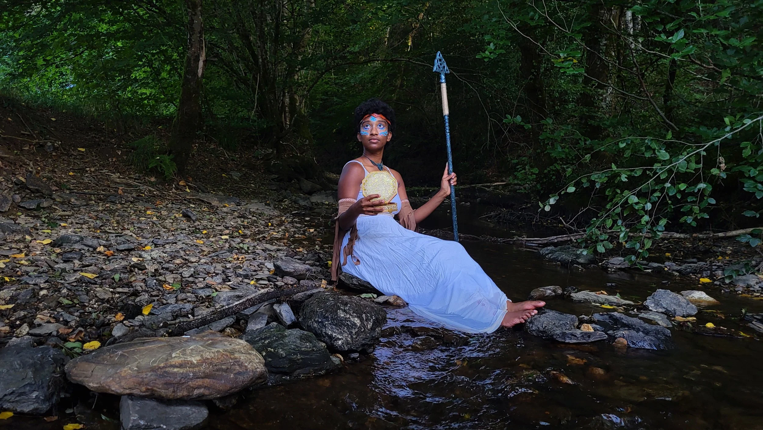 Mermaid Andrina as a Wild Water Goddess sitting in a river and holding a music box wearing warrior makeup