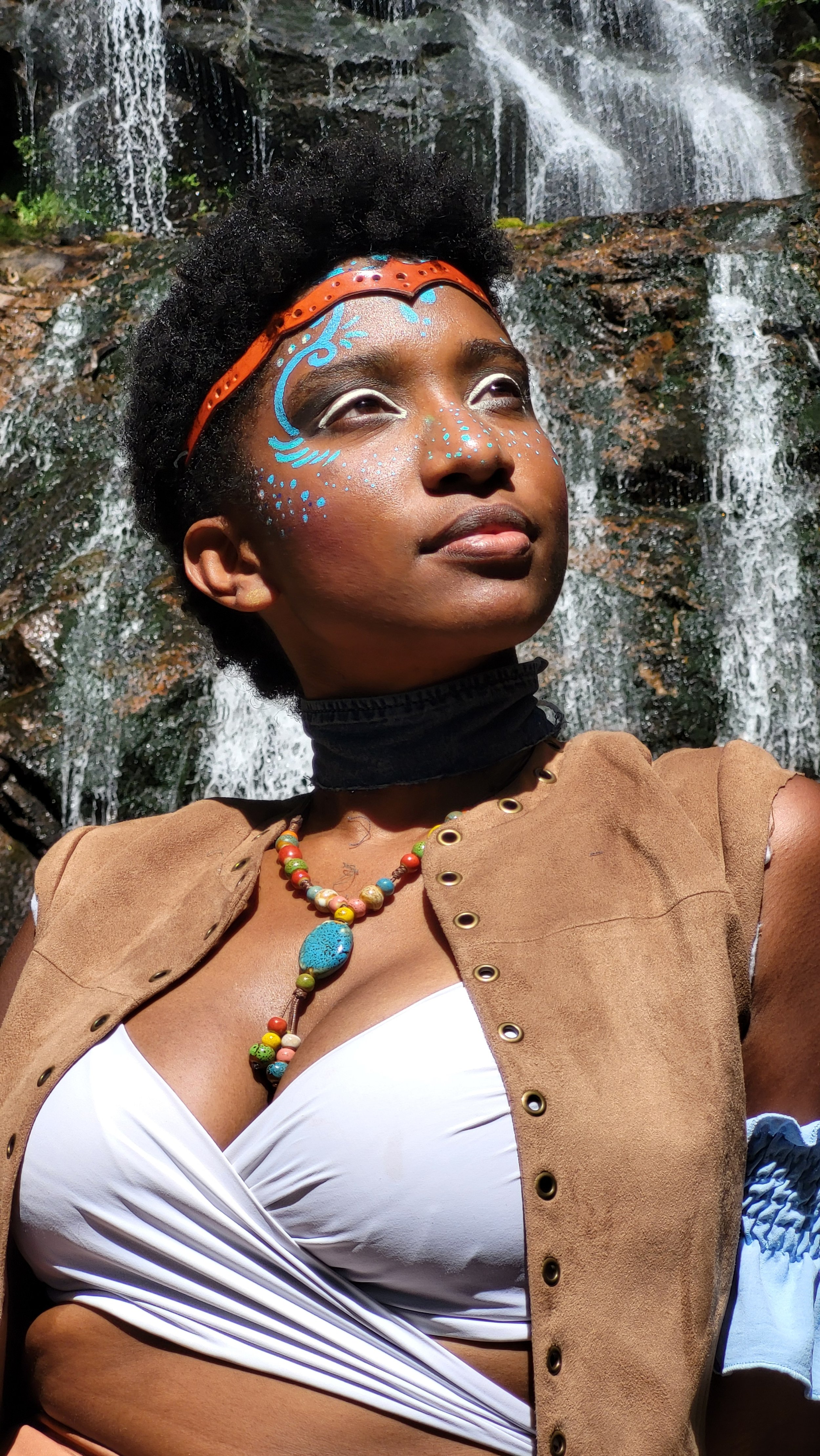 Mermaid Andrina with brown skin and curly black hair, wearing face paint with blue and white designs, a colorful beaded necklace, a brown vest, and a white top, sitting in front of a waterfall.