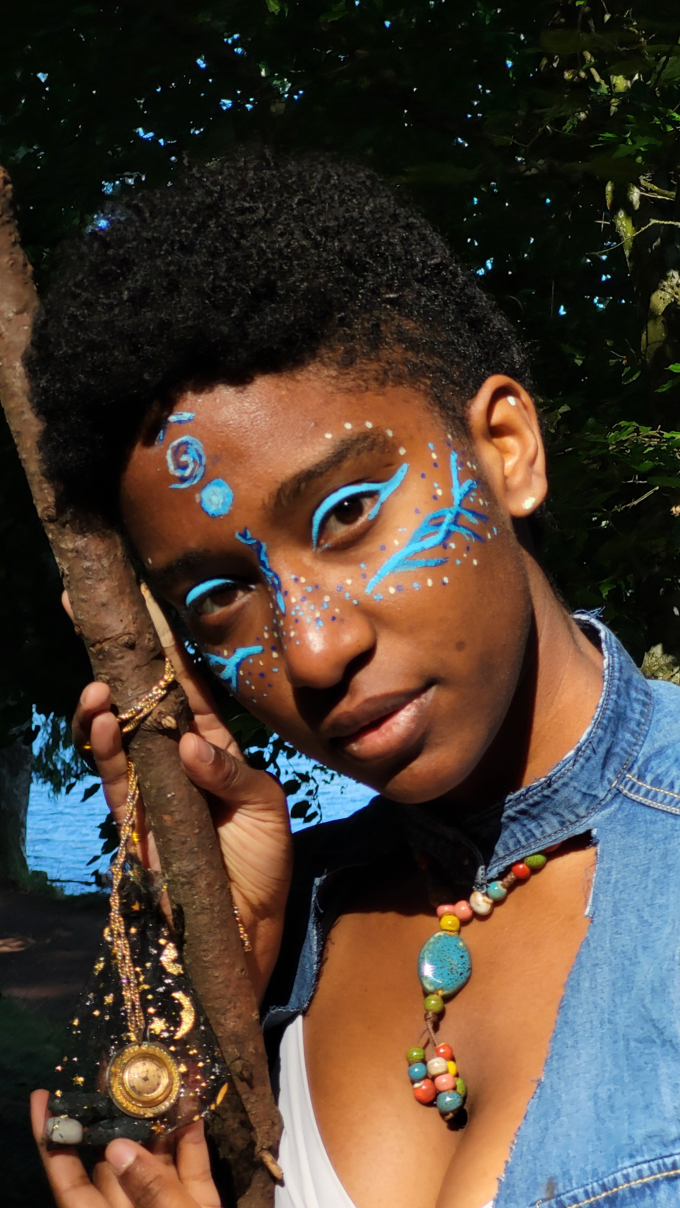 Mermaid Andrina in blue and white, holding a branch, wearing colorful beaded necklaces, and a watch with a black strap, outdoors in front of trees and water.