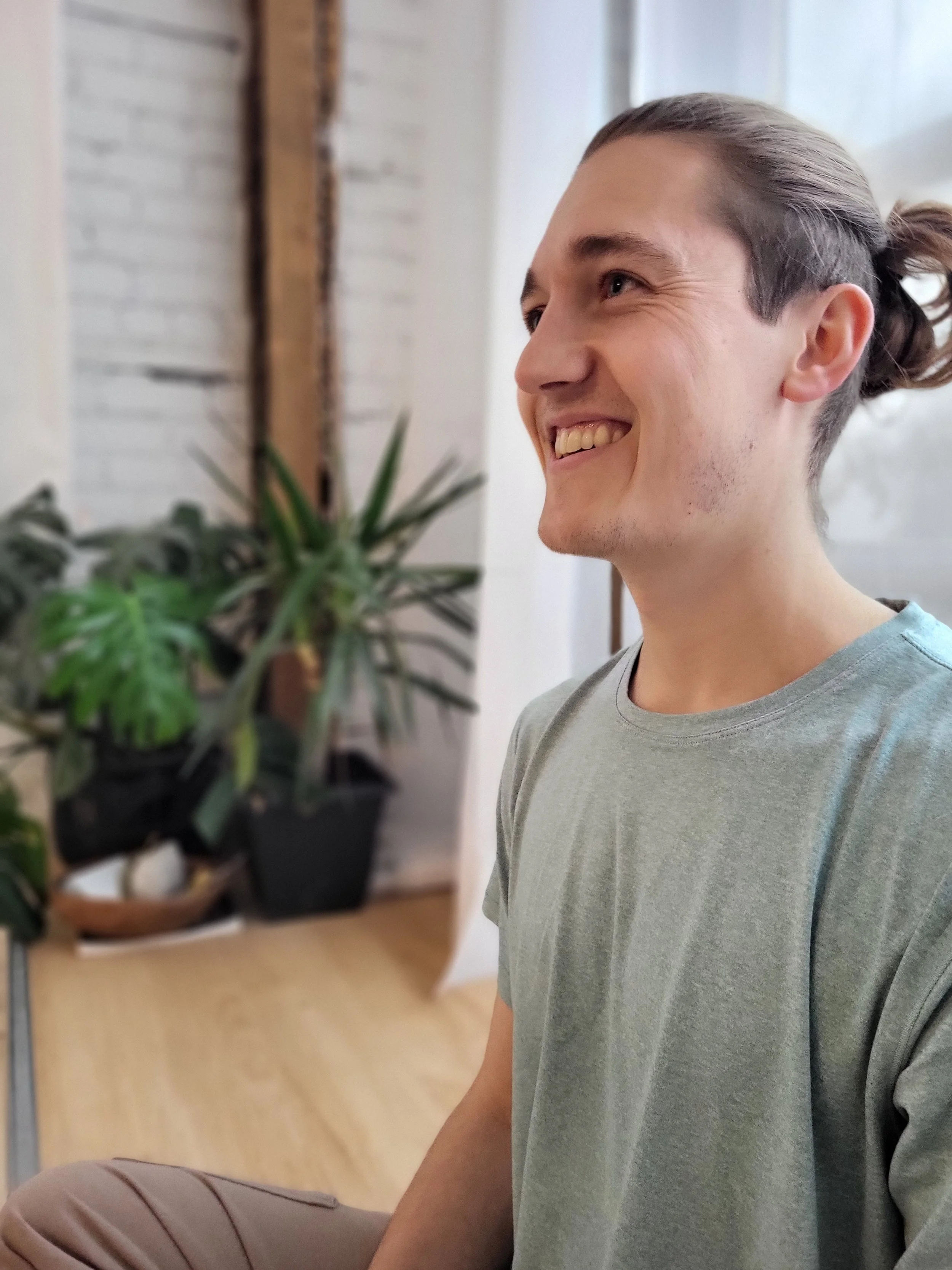 A young man with long hair tied back, smiling, sitting indoors near a potted plant with green leaves.