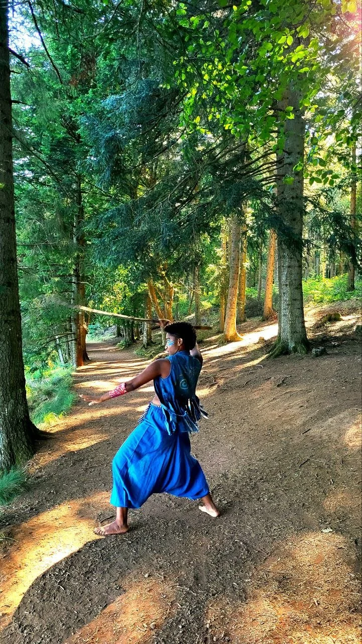Mermaid Andrina dressed in traditional blue clothing practicing martial arts with a sword in a lush green forest.