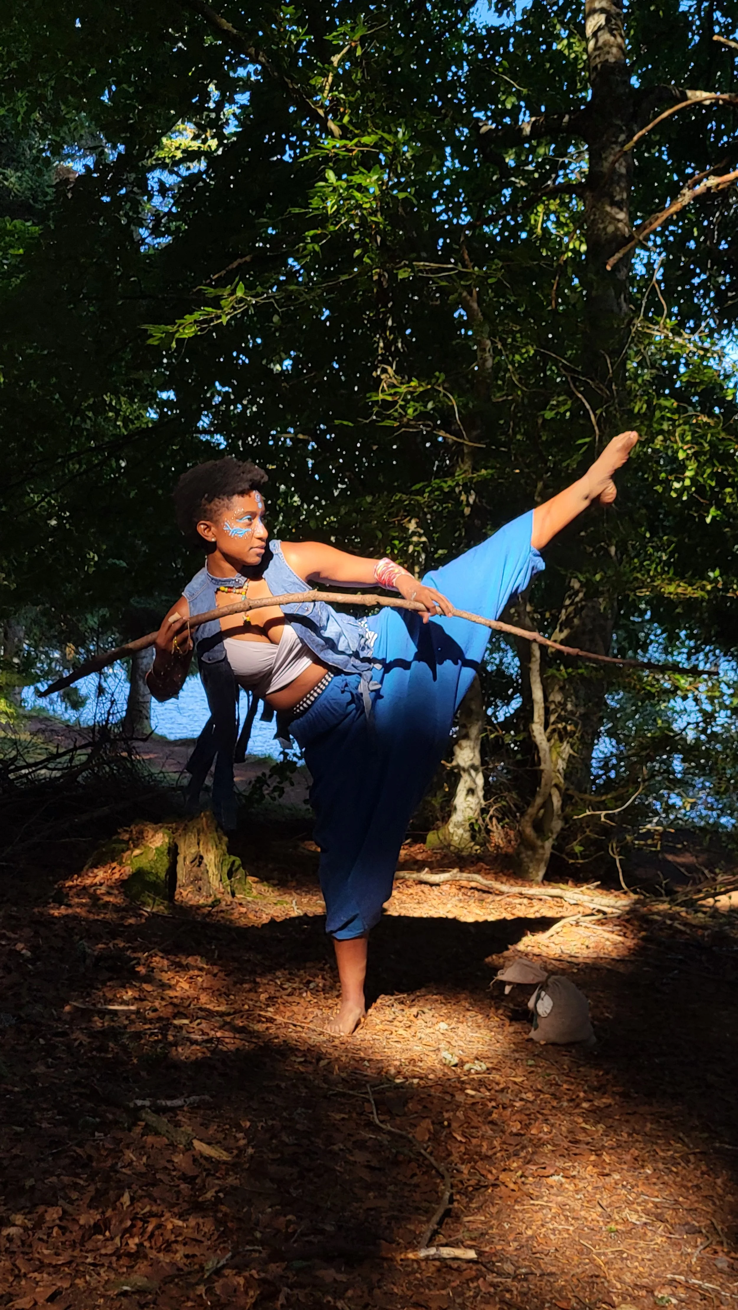 Mermaid Warrior Andrina in blue performs a high kick in a forested area during daylight, holding a stick and with face paint and accessories.