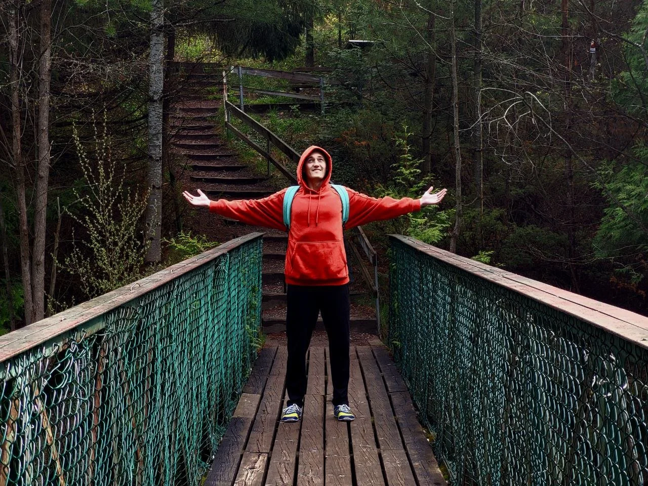 A person in a red hoodie and black pants standing on a wooden bridge with arms spread wide, surrounded by a forest with trees and greenery.