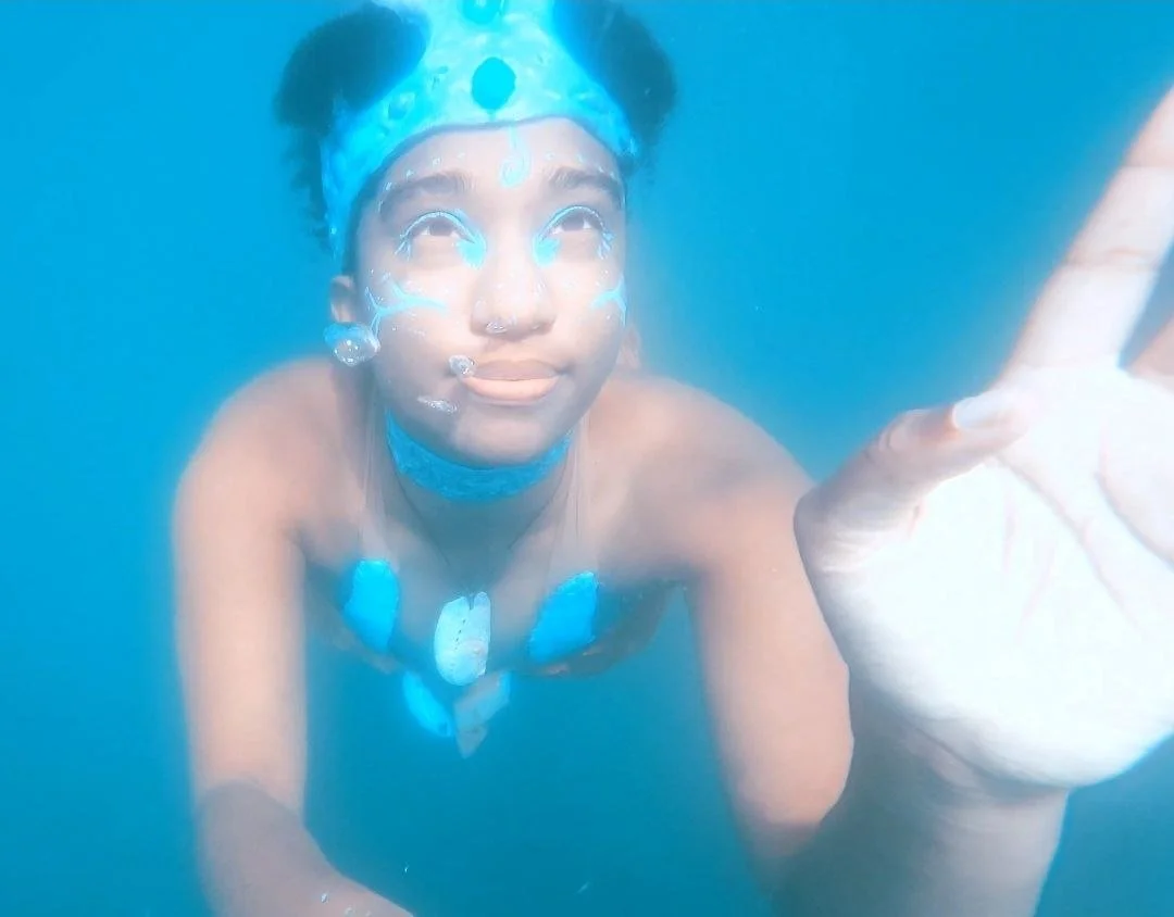 Mermaid Andrina, the humming mer (mermaid warrior), is just at the surface of the water with her tail behind her. Her face is half out of the water and her left hand reaches with fingers just out of the water towards the camera. The forest is behind.