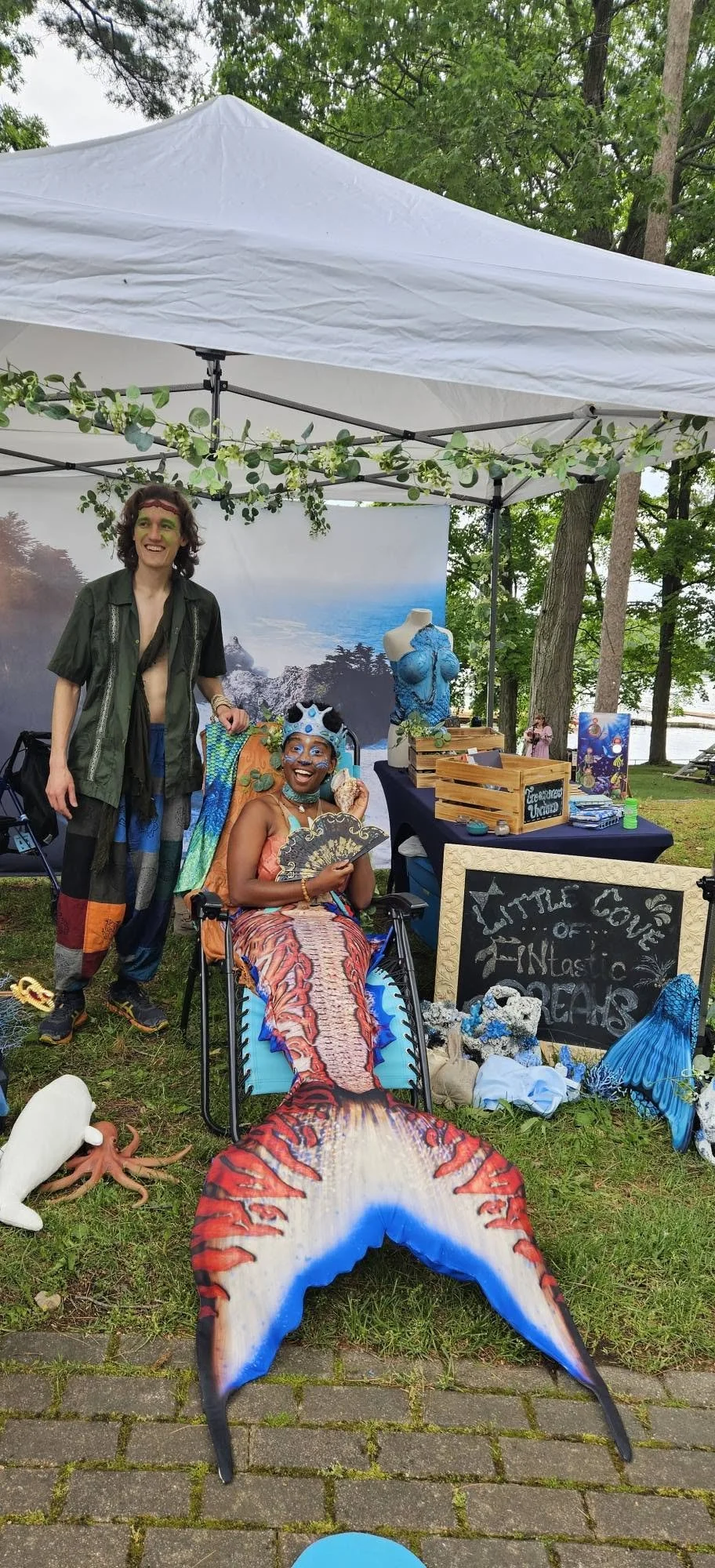 Two people in colorful costumes at an outdoor market booth decorated with ocean and mermaid-themed items, including a woman dressed as a mermaid with a scale-patterned tail and a man with face paint, standing under a white canopy with green foliage decorations.