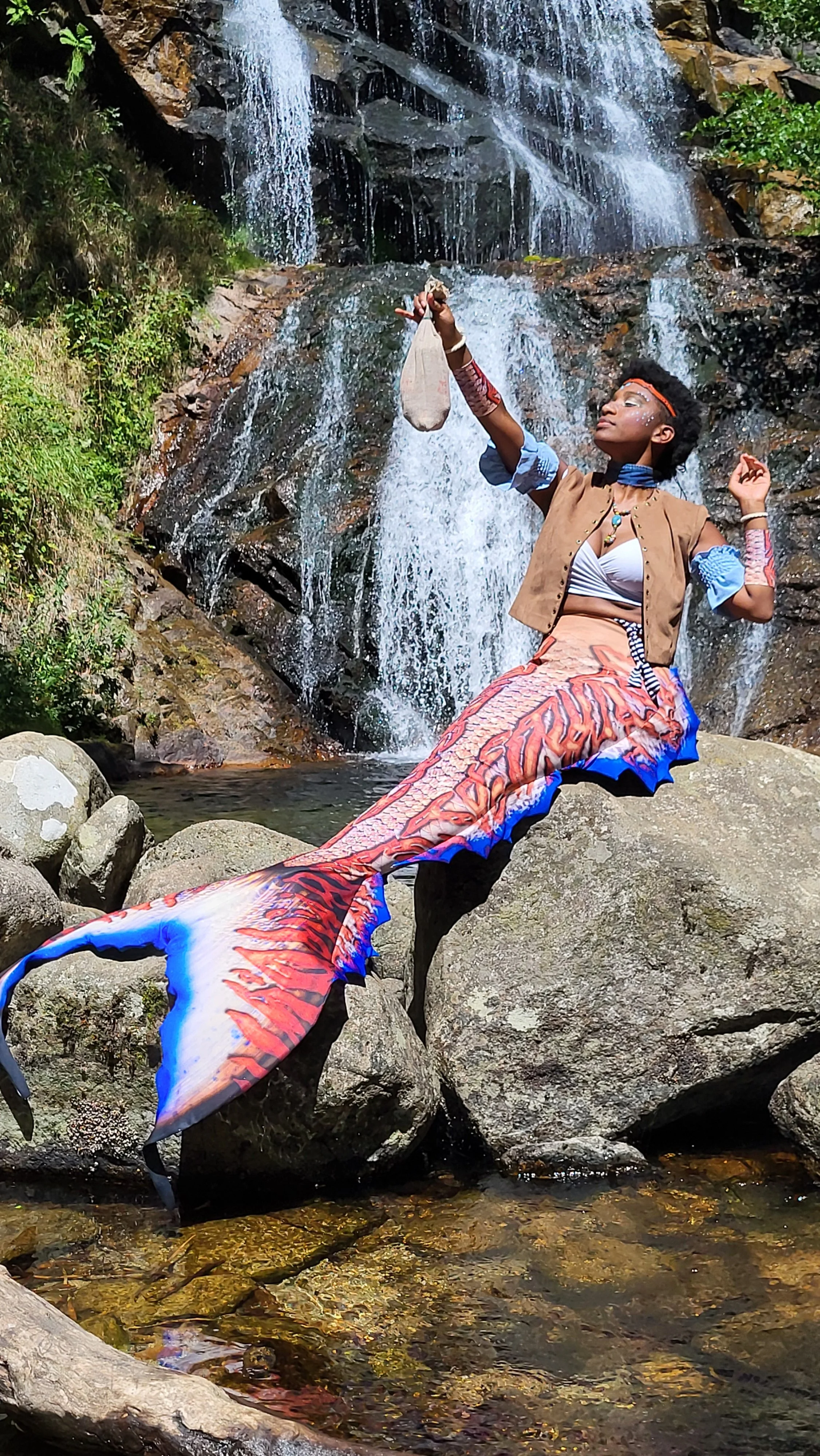 Mermaid Andrina sitting on rocks in a stream with a waterfall in the background. The individual holds a small bag in one hand and has decorative body art and accessories.