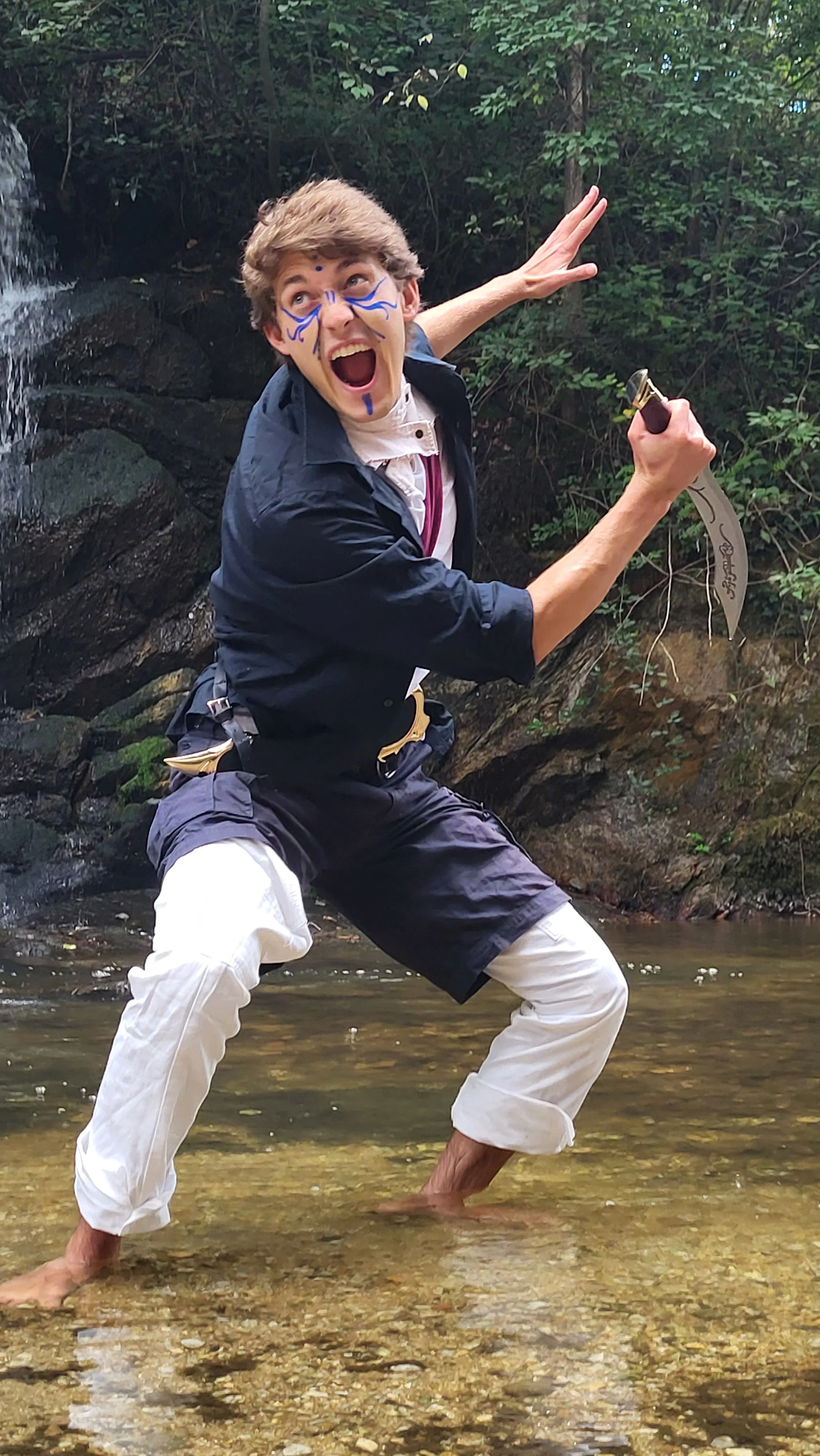 Octony in traditional martial arts uniform, posing in a creek with one foot in the water, surrounded by trees and rocks.